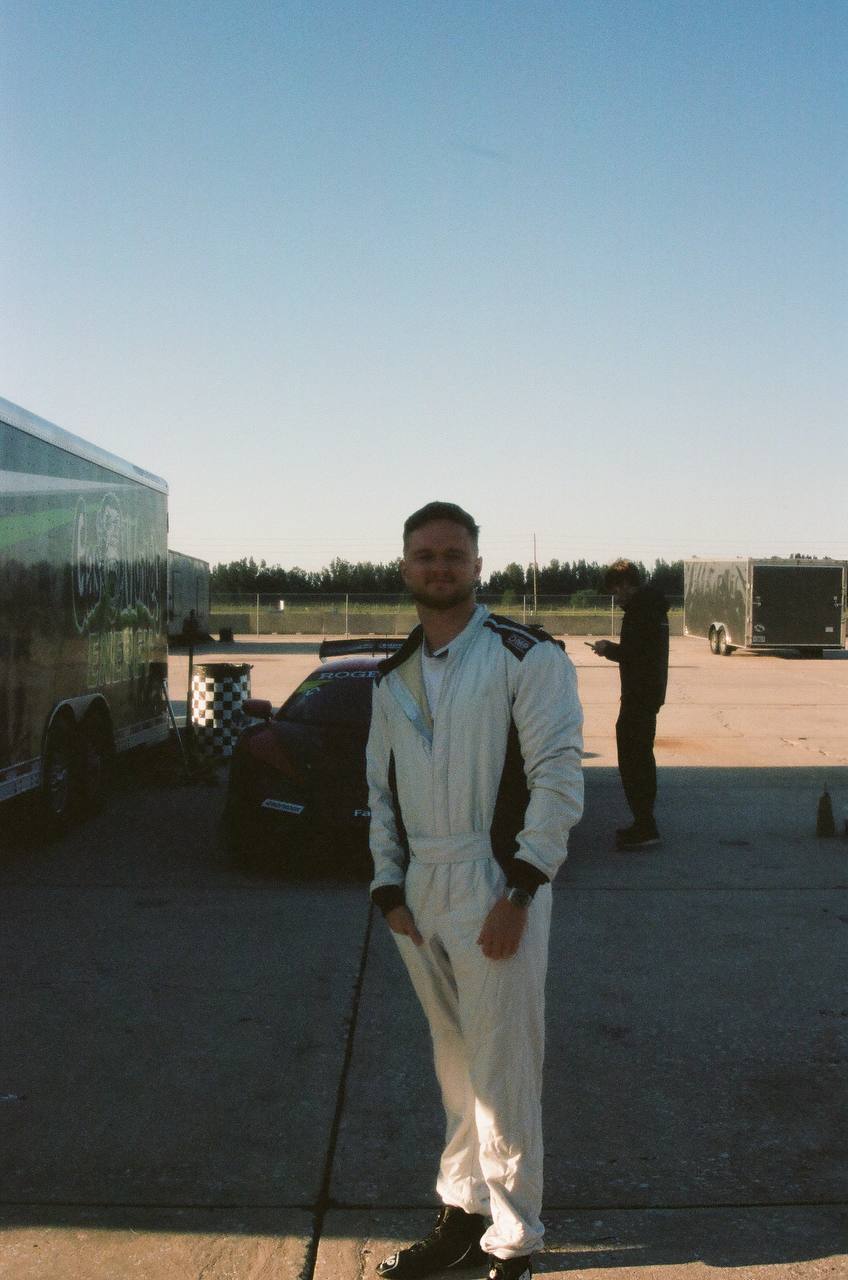 Dawson Gant in a full racing suit at the track with a race car behind him
